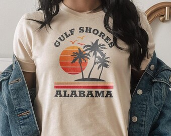 May include: A beige t-shirt with a graphic print of palm trees and a sunset. The text "GULF SHORES ALABAMA" is printed on the shirt.