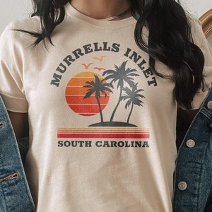 May include: A beige t-shirt with a graphic design of palm trees and a sunset. The text "Murrells Inlet South Carolina" is printed on the shirt.