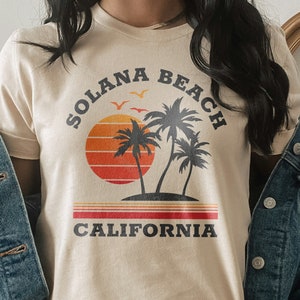May include: A beige t-shirt with a retro graphic design featuring palm trees, a sunset, and the text "Solana Beach California".