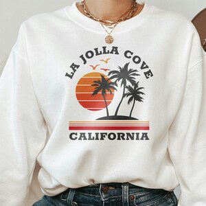 May include: White sweatshirt with a graphic print of a sunset, palm trees, and the text "La Jolla Cove California".
