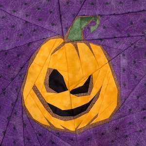 Pode incluir: Uma abóbora feita com peças de papel com uma cara assustadora, nas cores laranja, marrom e preto, sobre um fundo roxo com teias de aranha pretas.
