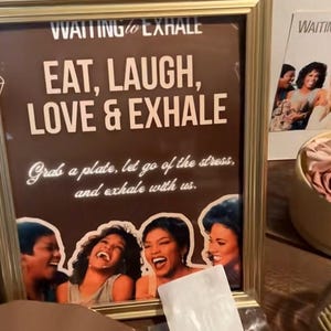 May include: A framed sign with the text "EAT, LAUGH, LOVE & EXHALE" in a brown background. The sign also includes the text "Grab a plate, let go of the stress, and exhale with us." and images of laughing women.
