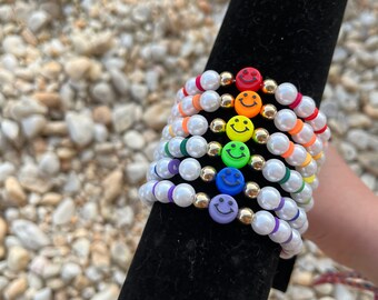 May include: A stack of colorful beaded bracelets with smiley face charms. The bracelets feature white faux pearls, gold beads, and smiley face charms in rainbow colors. The bracelets are displayed on a black velvet stand.
