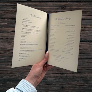May include: A wedding program, open to display the ceremony details and wedding party names. The cream-coloured pages are bound with a dark blue spine. The program is held against a dark wood background, with text in a classic font.