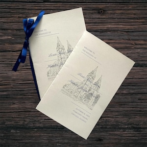 May include: Two cream wedding programs tied with a blue ribbon. The programs display a church illustration and text: "Welcome to the Wedding of Lexie Rose Love and Hunter Joseph Hearst" and the date October 18, 2025.