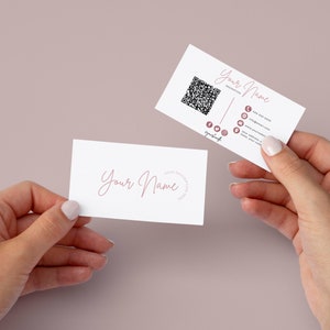 May include: Two white business cards with pink text. The top card has a QR code and social media icons. The text on the card reads "Your Name" and includes contact information. The bottom card has the text "Your Name" in cursive.