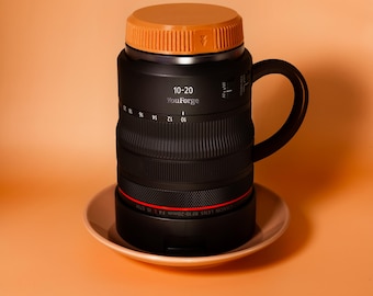 May include: A black mug shaped like a camera lens with a handle, featuring the text 10-20 and YouForge on the side. The mug has a bright orange lid and sits on a matching orange saucer. The lens barrel design includes white focal length markings.