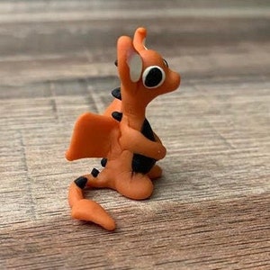 May include: A small orange and black dragon figurine with white eyes. The dragon is holding its wings close to its body.