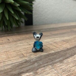 Puede incluir: Una pequeña figurita pintada a mano de un animal blanco y negro sosteniendo un objeto azul en forma de corazón. La figurita está hecha de arcilla polimérica y tiene un estilo caprichoso y de dibujos animados.