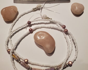 Puede incluir: Cuentas de cintura blancas con detalles en rosa y plata se disponen con piedras rosas y un cristal de selenita blanco. Las cuentas de cintura tienen unas cuentas rectangulares más grandes con un patrón morado y marrón.