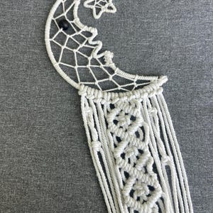 May include: White macrame dream catcher featuring a crescent moon shape with a star accent. The dream catcher has a woven web design within the moon and hanging knotted cords. A black bead is attached to the moon.
