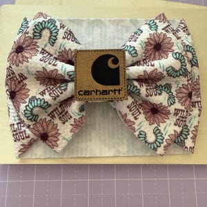 May include: A floral headband with a Carhartt logo patch. The fabric features a pattern of pink and teal flowers with the words "Wild Soul". The headband is on a wooden surface.
