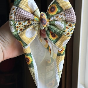 Könnte beinhalten: Eine große, handgefertigte Haarschleife mit einem Patchwork-Design mit Sonnenblumen in Gelb, Grün und Braun. Die Schleife wird hochgehalten und zeigt ihren strukturierten Stoff und das detaillierte Sonnenblumenmuster. Haarschmuck.