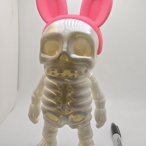 Può includere: Una figura scheletrica bianca traslucida con una fascia per orecchie da coniglio rosa. La figura ha una testa a forma di teschio e un design a gabbia toracica. Un pennarello nero è accanto alla statuina.