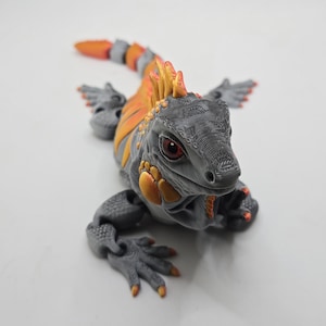 May include: A 3D printed articulated iguana figurine featuring a grey body with vibrant orange and yellow accents along the spine, head, and tail. The lizard has detailed textured skin, dark eyes, and segmented limbs that allow for movement.