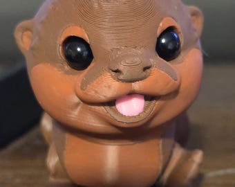 May include: A 3D-printed otter figure in brown and orange, with black eyes and a pink tongue. The text "Realistic 3 Color" is at the bottom. The figure is a cute, cartoon-like design.