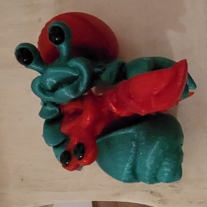 May include: A 3D printed toy of a red and teal hermit crab. The crab has large eyes and a detailed shell.