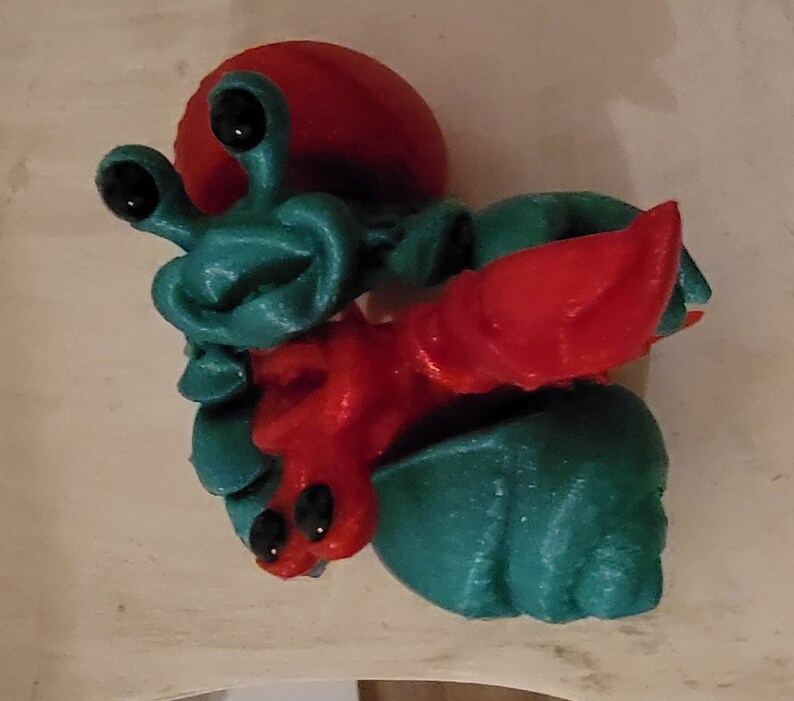 May include: A 3D printed toy of a red and teal hermit crab. The crab has large eyes and a detailed shell.