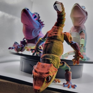 Könnte beinhalten: Drei artikulierte Gecko-Figuren, eine ist rot, orange und gelb, eine ist lila und blau und eine ist rosa. Die Figuren sind spielerisch in Szene gesetzt.
