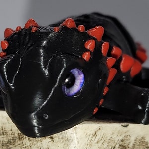 Peut inclure: Un petit dragon noir imprimé en 3D avec des pointes rouges et des yeux violets. Le dragon est couché sur le ventre, la tête tournée sur le côté.