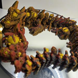 May include: A detailed, segmented sculpture of a creature with a serpentine form. The sculpture features a mix of brown, red, and yellow hues, with intricate detailing. The creature is displayed on a reflective surface.