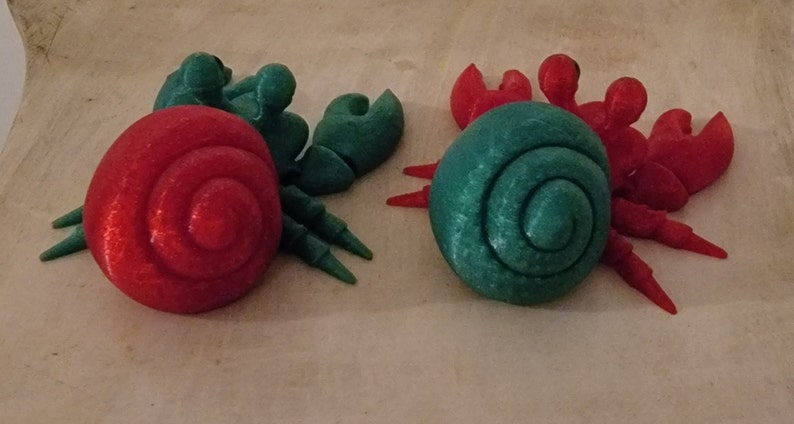 May include: Two 3D printed hermit crabs, one with a red shell and green legs, the other with a green shell and red legs.