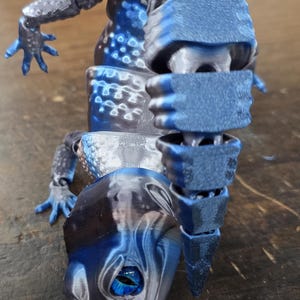 May include: A 3D printed blue and gray lizard figurine with articulated limbs. The lizard has a segmented body and a blue eye.