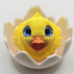 May include: A yellow chick with blue eyes and an orange beak emerging from a white eggshell. The chick has textured feathers and is centered in the frame. The eggshell has a light pink interior.