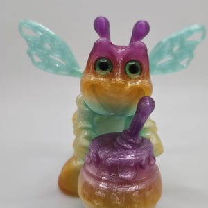 May include: A vibrant 3D-printed bee-like figure with a honey pot. The figure has a gradient of colours, including yellow, orange, purple, and green. The bee has large green eyes and translucent wings. The honey pot is also multi-coloured.