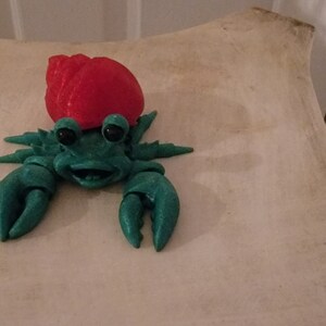 May include: A green and red plastic toy hermit crab with a smiling face. The crab is wearing a red shell and has large, black eyes.