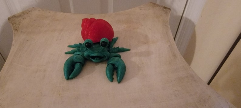 May include: A green and red plastic toy hermit crab with a smiling face. The crab is wearing a red shell and has large, black eyes.