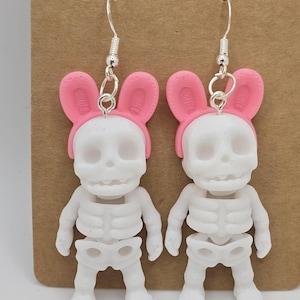 Può includere: Un paio di orecchini originali con figure di scheletri bianchi che indossano fasce per le orecchie da coniglio rosa. Gli orecchini hanno fili per orecchie a gancio di pesce color argento e sono esposti su uno sfondo marrone. Le figure degli scheletri hanno gabbie toraciche e arti dettagliati.