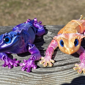 Peut inclure: Deux petites figurines d'gecko articulées et colorées. L'une est violette et bleue, l'autre est orange et rose.