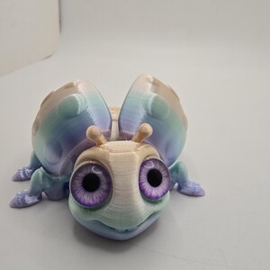 May include: A 3D-printed, whimsical insect figurine with large, purple-eyed, and a smiling face. The wings and body feature a gradient of pastel colors, including blue, green, and tan. The insect has small antennae and six legs.