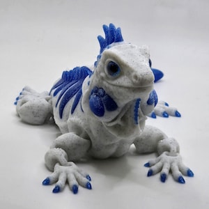 Peut inclure: Une figurine d'iguane articulée imprimée en 3D avec un corps blanc moucheté de noir et des accents bleu vif sur la crête, les membres, les griffes et les détails du visage. Le lézard présente une surface texturée et un design segmenté.