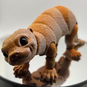 May include: A detailed, 3D-printed figurine of a stylized, armadillo-like creature. The figure is primarily brown with lighter tan accents, featuring segmented body sections and large, expressive eyes. It is posed on a small, textured base.