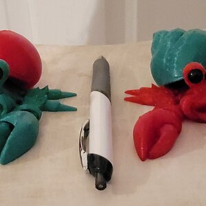 May include: Two 3D printed hermit crabs, one with a red shell and green body, the other with a green shell and red body. Both have black eyes and a smiling face.