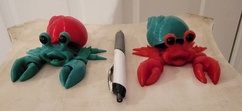 May include: Two 3D printed hermit crabs, one with a red shell and green body, the other with a green shell and red body. Both have black eyes and a smiling face.