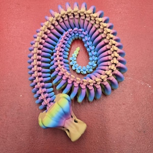 Op de afbeelding: Een kleurrijke, meerkleurige, plastic, 3D-geprinte, gesegmenteerde, slangachtige ketting met een regenboogkleurige gradiënt. De ketting is opgerold in een spiraalvorm.