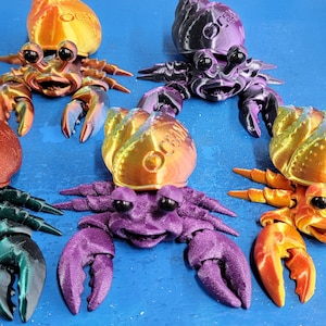 May include: Five colorful, 3D printed hermit crabs with iridescent paint. Each crab has a different color, including orange, purple, green, black, and red. The crabs are all facing forward with their claws outstretched.