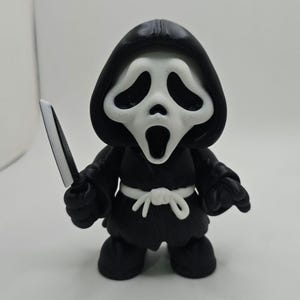 May include: A small figurine of a character wearing a black hooded robe with a white waist tie and a white mask featuring a long, distorted mouth and eye openings. The character holds a small, silver-colored knife in its right hand.