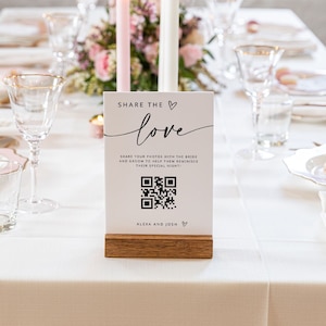 Könnte beinhalten: Eine weiße Karte mit schwarzem Text, auf der "Teile die Liebe" steht und ein QR-Code abgebildet ist. Die Karte steht auf einem Holzständer und liegt auf einer weißen Tischdecke mit Kerzen und Blumen.