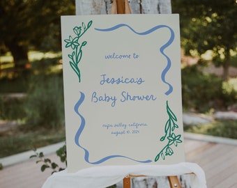 May include: A wooden easel displays a sign for Jessica's Baby Shower. The sign is cream-colored with blue and green floral accents and text that reads "welcome to Jessica's Baby Shower" and event details. A white cloth drapes the easel.