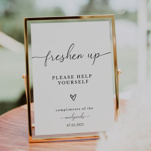 May include: A gold-framed sign with the text "freshen up" in cursive, followed by "PLEASE HELP YOURSELF" in all caps. Below the text is a small heart and the words "compliments of the newlyweds" in cursive. The date "07.03.2023" is printed below the cursive text.