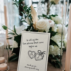 May include: A white card with black text and a hand-drawn illustration of two locket hearts with the letters "M" and "J" inside. The text reads "To all of the love stories that came before us. Thank you for showing us the way."
