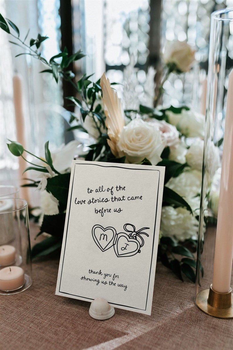 May include: A white card with black text and a hand-drawn illustration of two locket hearts with the letters "M" and "J" inside. The text reads "To all of the love stories that came before us. Thank you for showing us the way."
