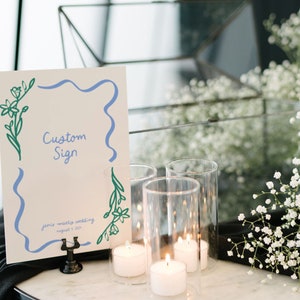 Op de afbeelding: Een wit bord met een blauwe en groene bloemenrand en de tekst "Custom Sign" en "Jamie + Matteo Wedding August 1, 2023". Het bord staat op een zwarte standaard en er zijn kaarsen en witte bloemen op de achtergrond.