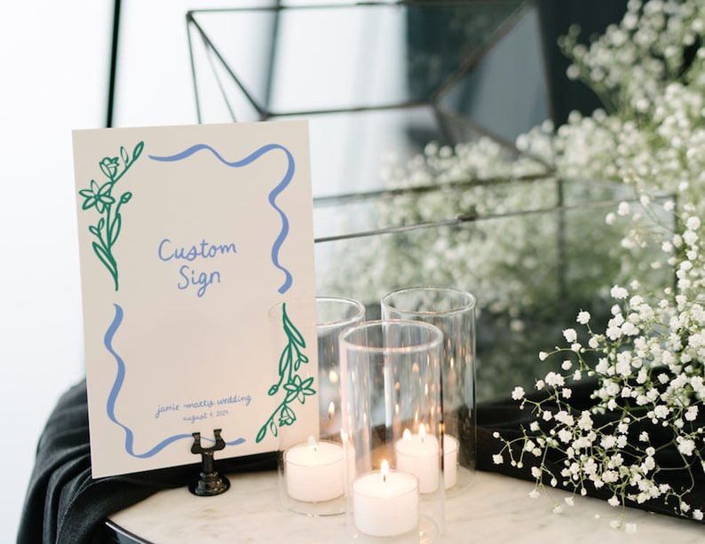 Op de afbeelding: Een wit bord met een blauwe en groene bloemenrand en de tekst "Custom Sign" en "Jamie + Matteo Wedding August 1, 2023". Het bord staat op een zwarte standaard en er zijn kaarsen en witte bloemen op de achtergrond.