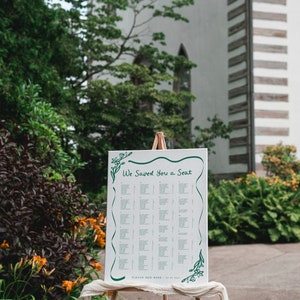May include: A white wedding seating chart with green floral accents on an easel. The chart is titled "We Saved You a Seat" and lists guest names and table assignments.