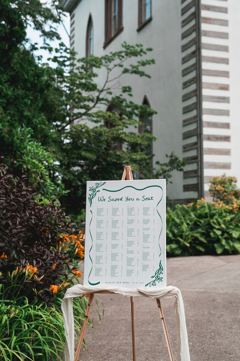 May include: A white wedding seating chart with green floral accents on an easel. The chart is titled "We Saved You a Seat" and lists guest names and table assignments.